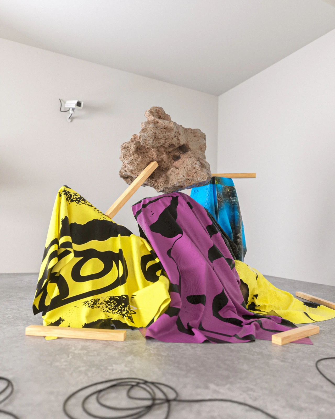3D installation in a white room combines a rough rock, wooden battens and yellow and magenta fabrics with black graphics beneath a surveillance camera.