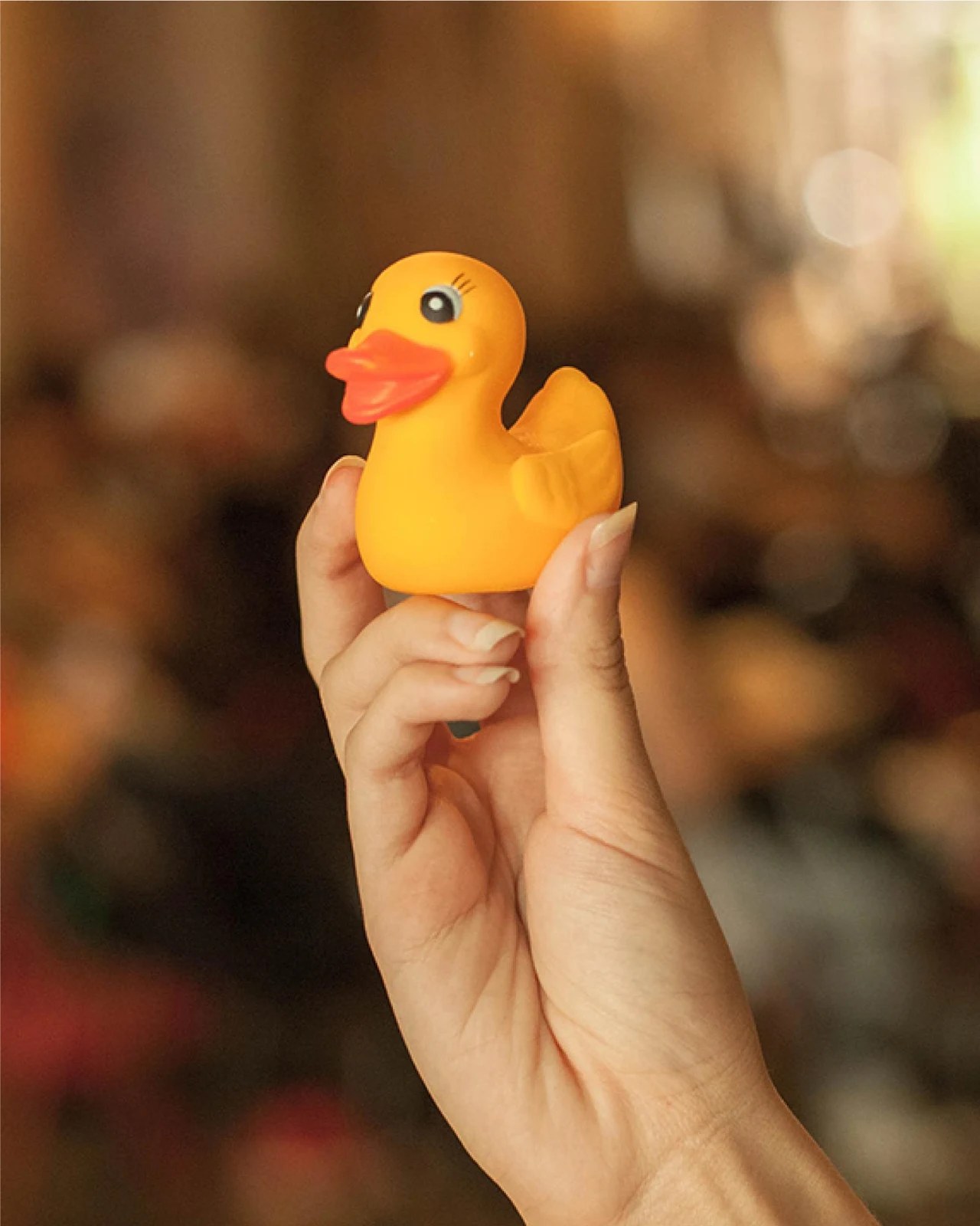 A hand raises a small yellow rubber duck in front of a blurred, warmly lit interior.