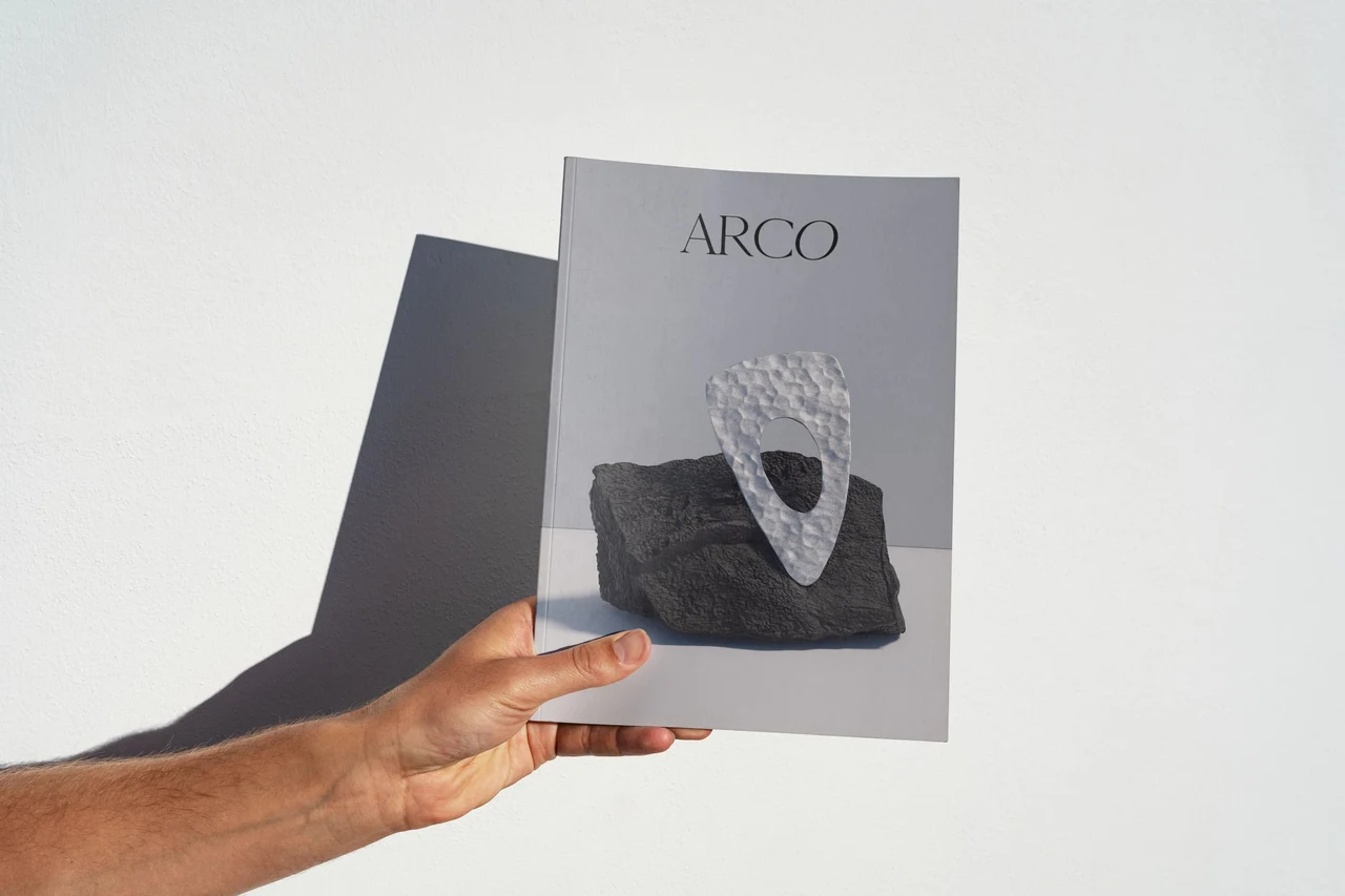 A hand holds an ARCO print featuring silver jewelry on dark rock, with side lighting casting a strong shadow on the wall.