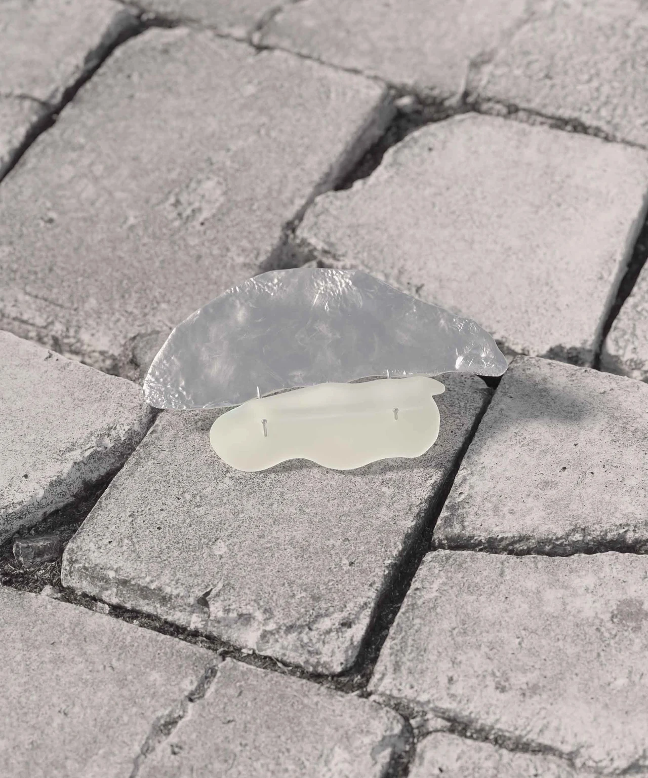 A jewelry piece combining a metallic organic shape and a pale one rests on gray paving stones.