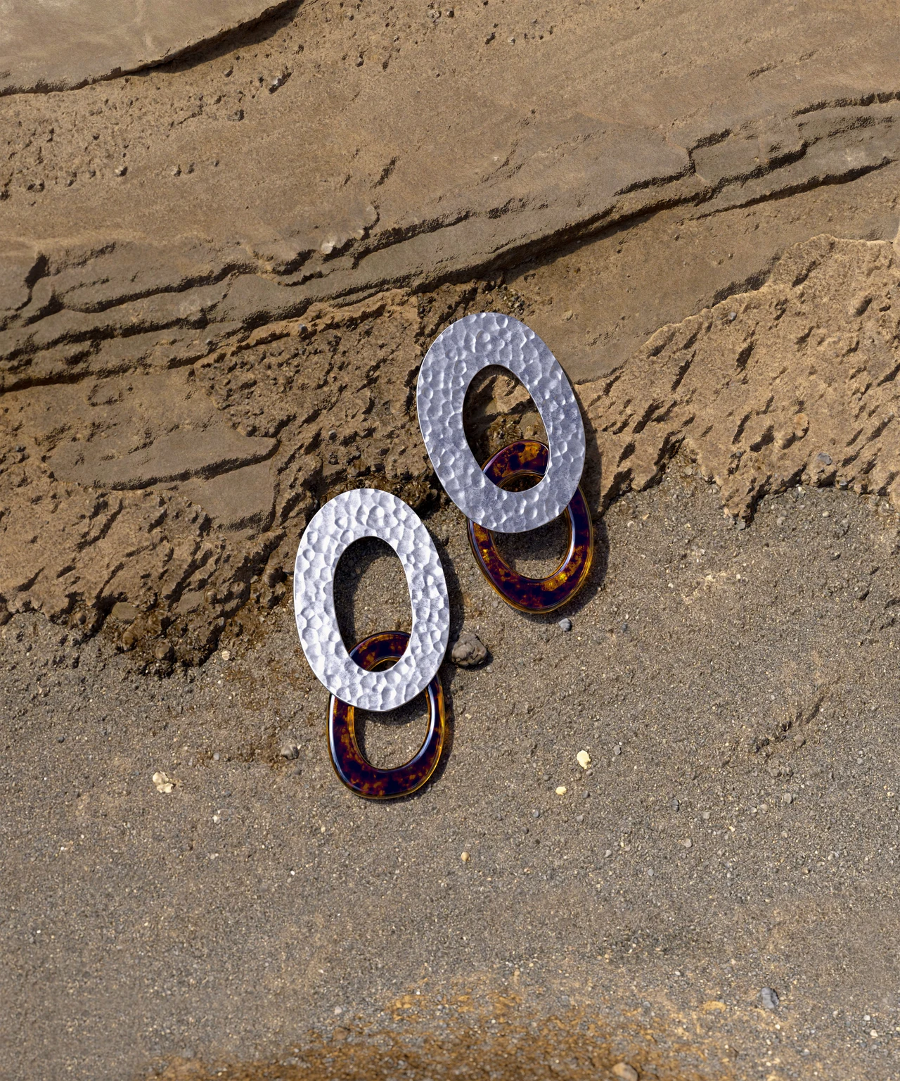 A pair of earrings with oval silver rings and golden loops rests on sand beside a layered stone wall.