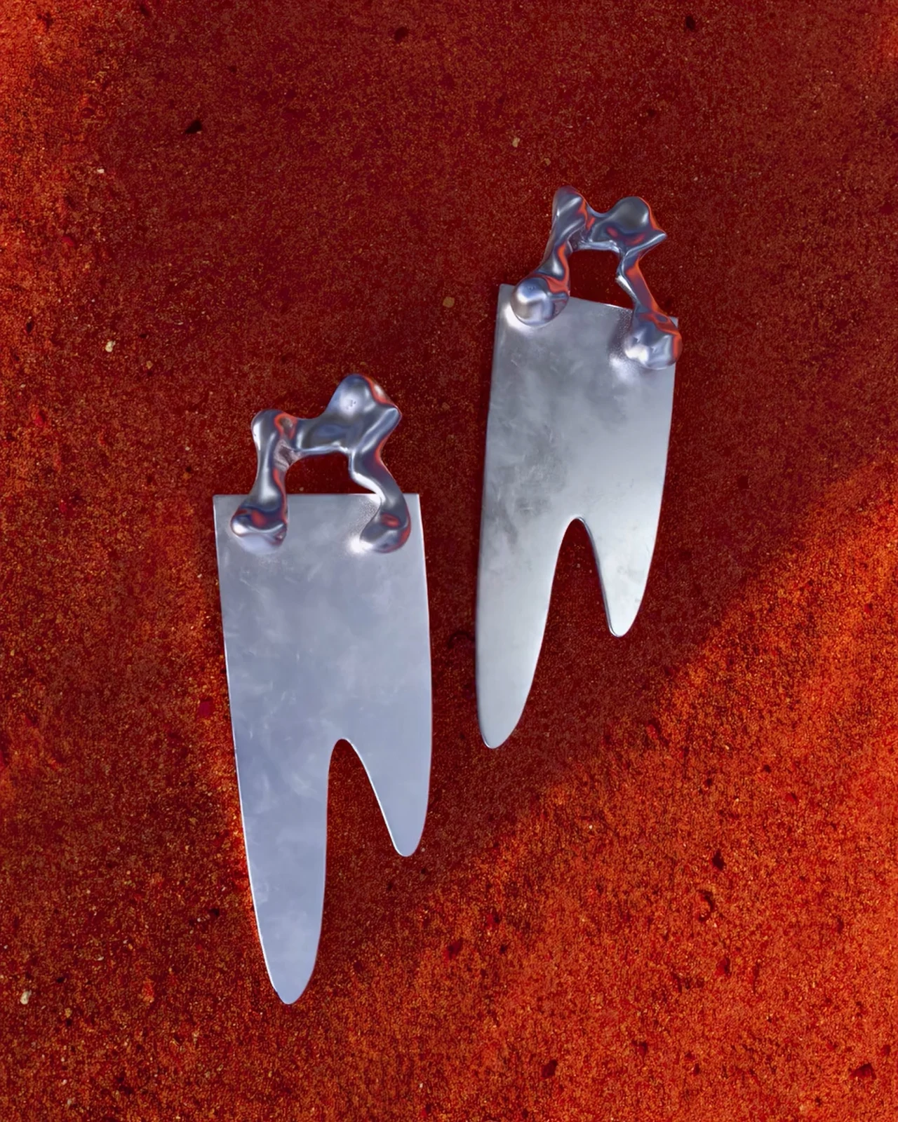 A pair of elongated silver earrings with organic cutouts and small upper elements rests on a red mineral surface.
