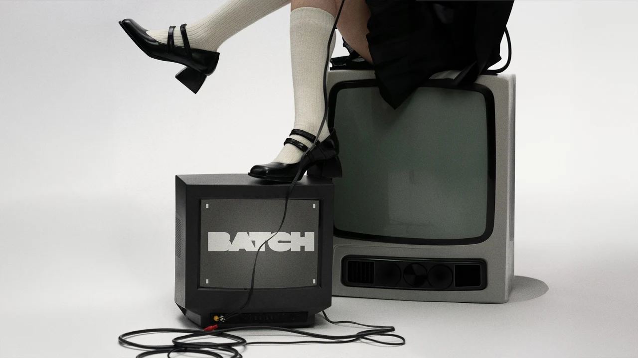 Studio photograph of a person's legs seated on CRT televisions, black shoes and cables on the floor; the smaller monitor displays BATCH.