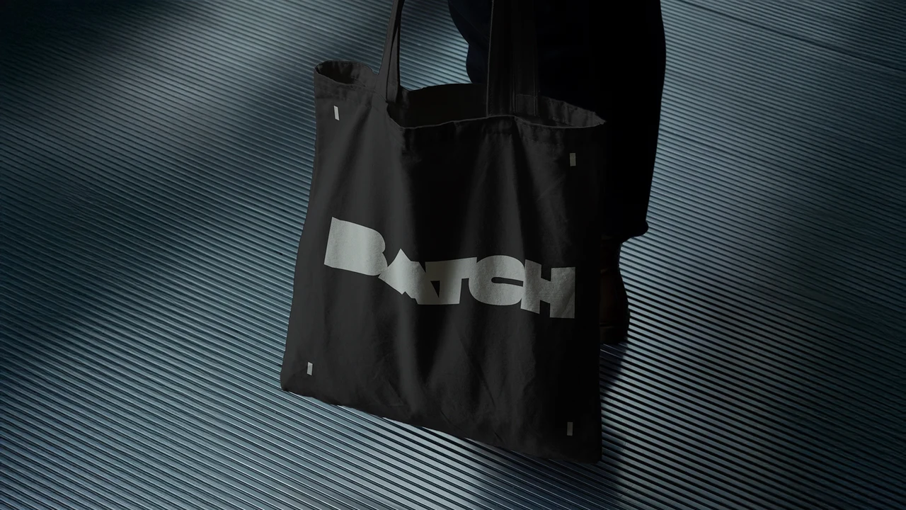 Black fabric tote bag with a pale BATCH wordmark photographed in low light against a dark diagonally striped background.