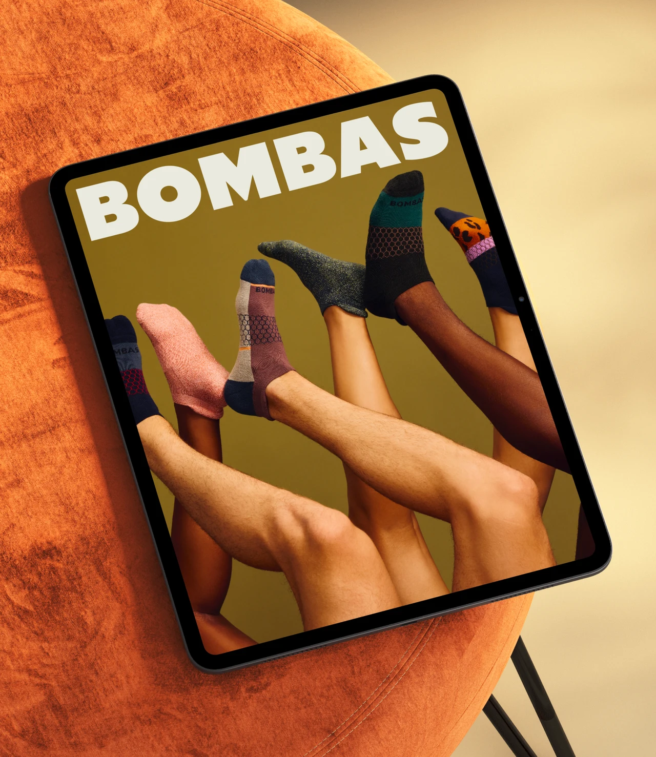 Tablet resting on an orange velvet chair, its screen showing the BOMBAS wordmark over legs in colorful socks against a mustard background.
