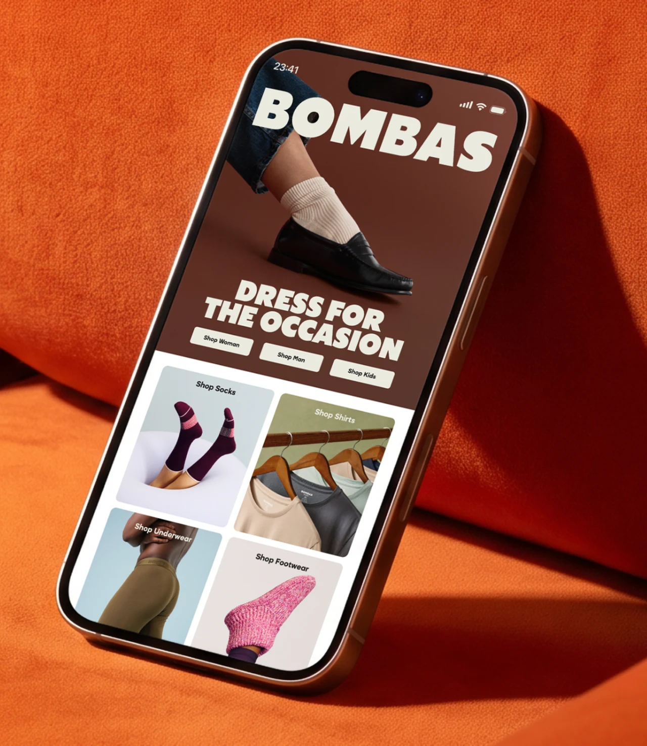 Smartphone leaning on orange velvet shows the Bombas site: the BOMBAS wordmark, a Dress for the Occasion banner with a white sock and black loafer, and tiles for socks, shirts, underwear and footwear.