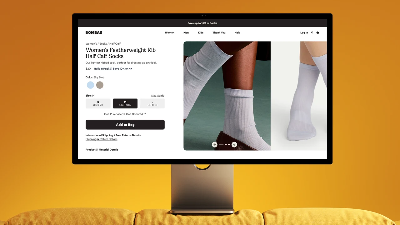 Desktop monitor on a yellow sofa displays a Bombas product page for Women's Featherweight Rib Half Calf Socks, with photos of pale blue ribbed socks.