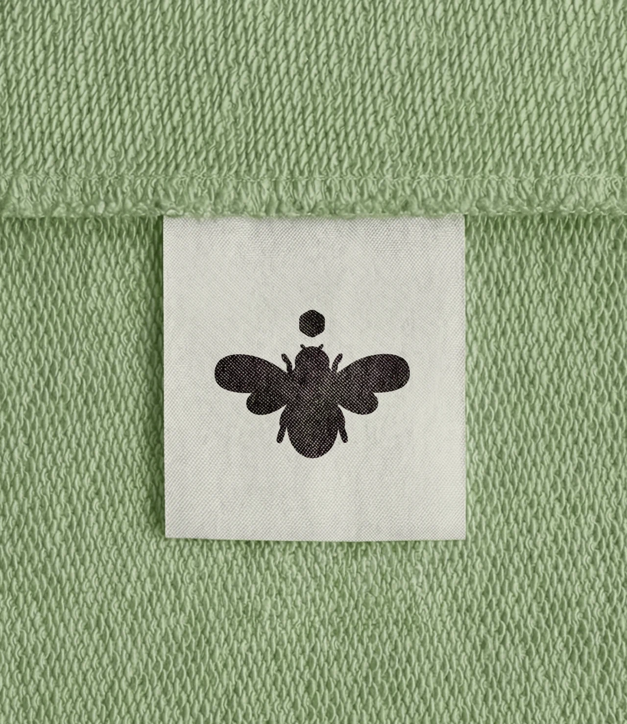 Close-up of a white woven label with a black bee icon stitched onto green terry fabric.
