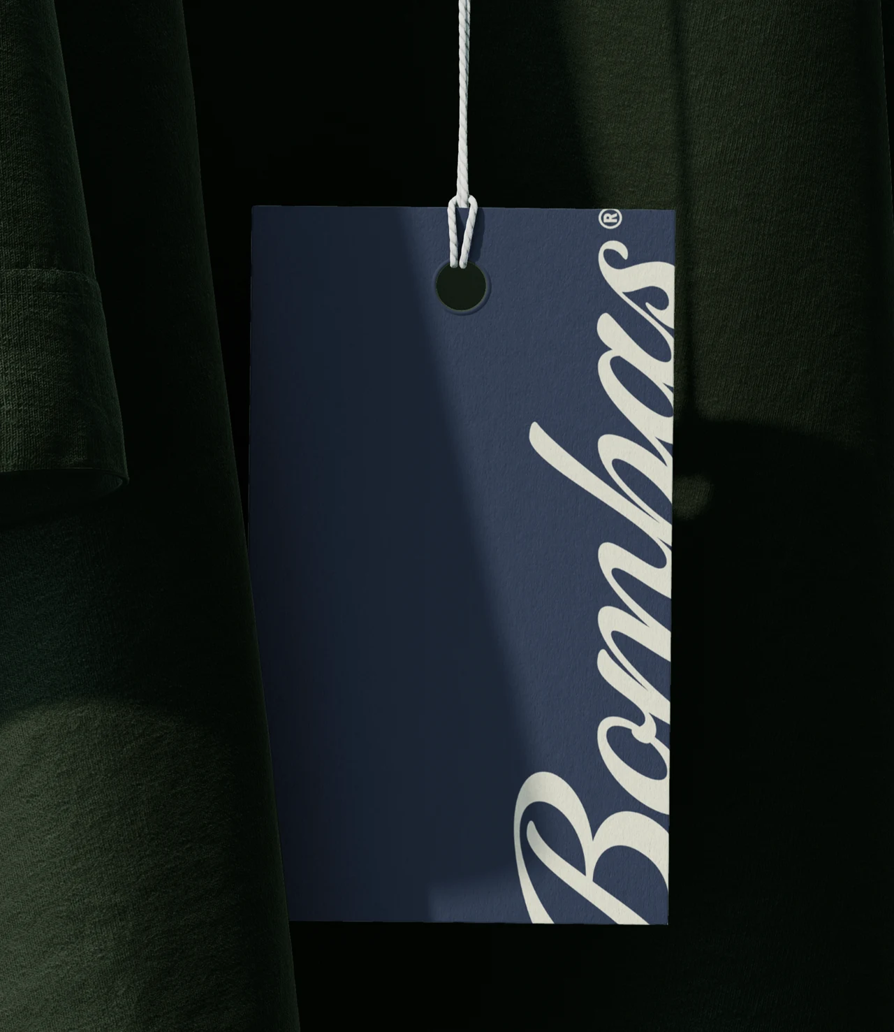 Navy hang tag with the cream script Bombas logo, hanging from a white string against dark green fabric.
