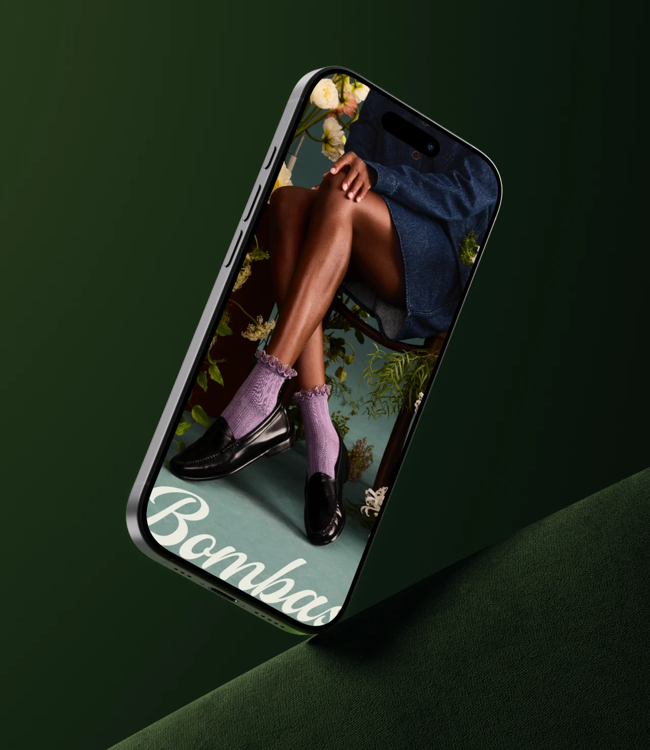 Smartphone on dark green velvet showing a photo of crossed legs in lilac ruffled socks and black loafers among flowers, with the script Bombas logo across the screen.