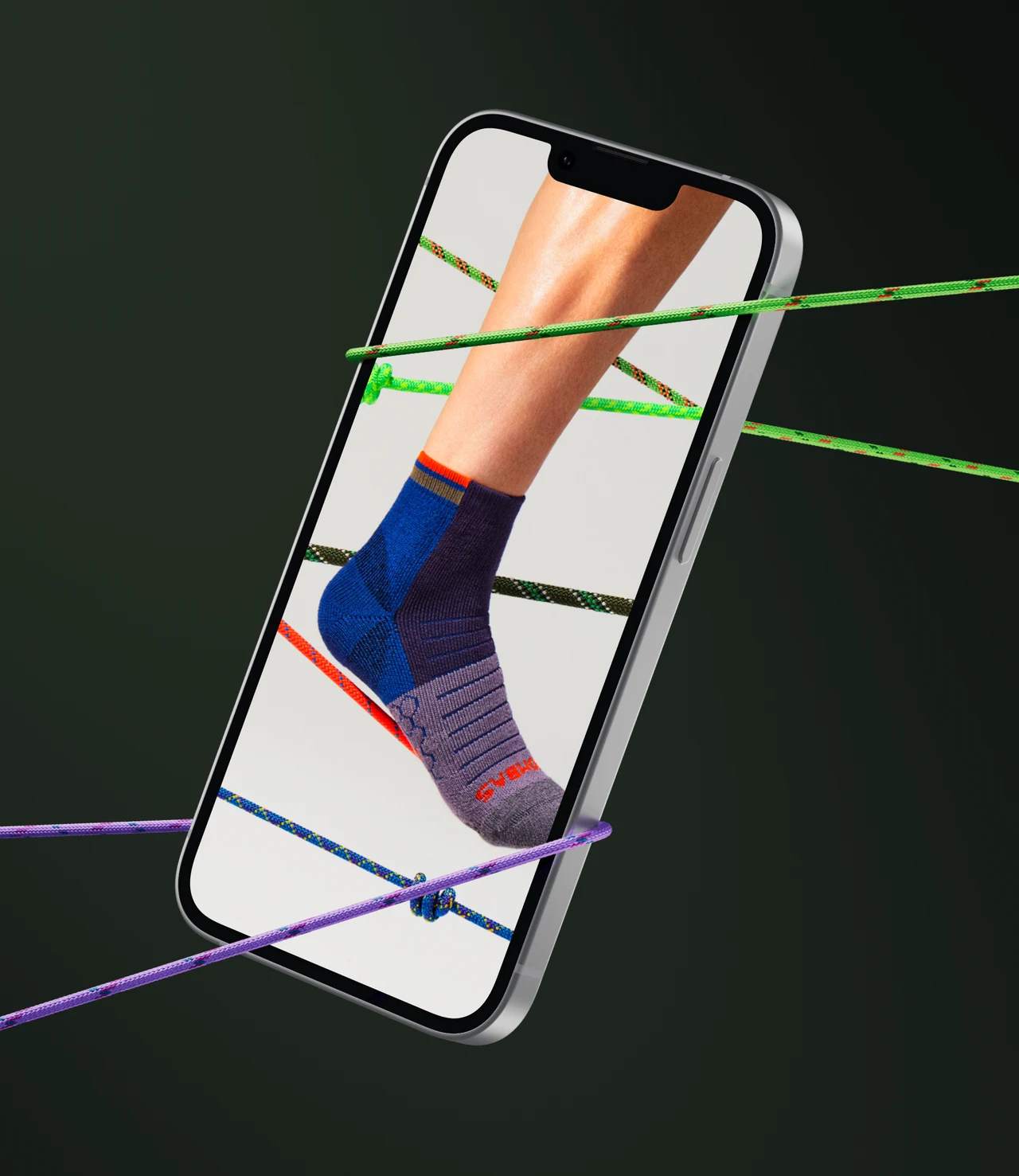 Smartphone suspended among colorful climbing ropes on a dark green background, its screen showing a foot in a blue and purple athletic ankle sock.