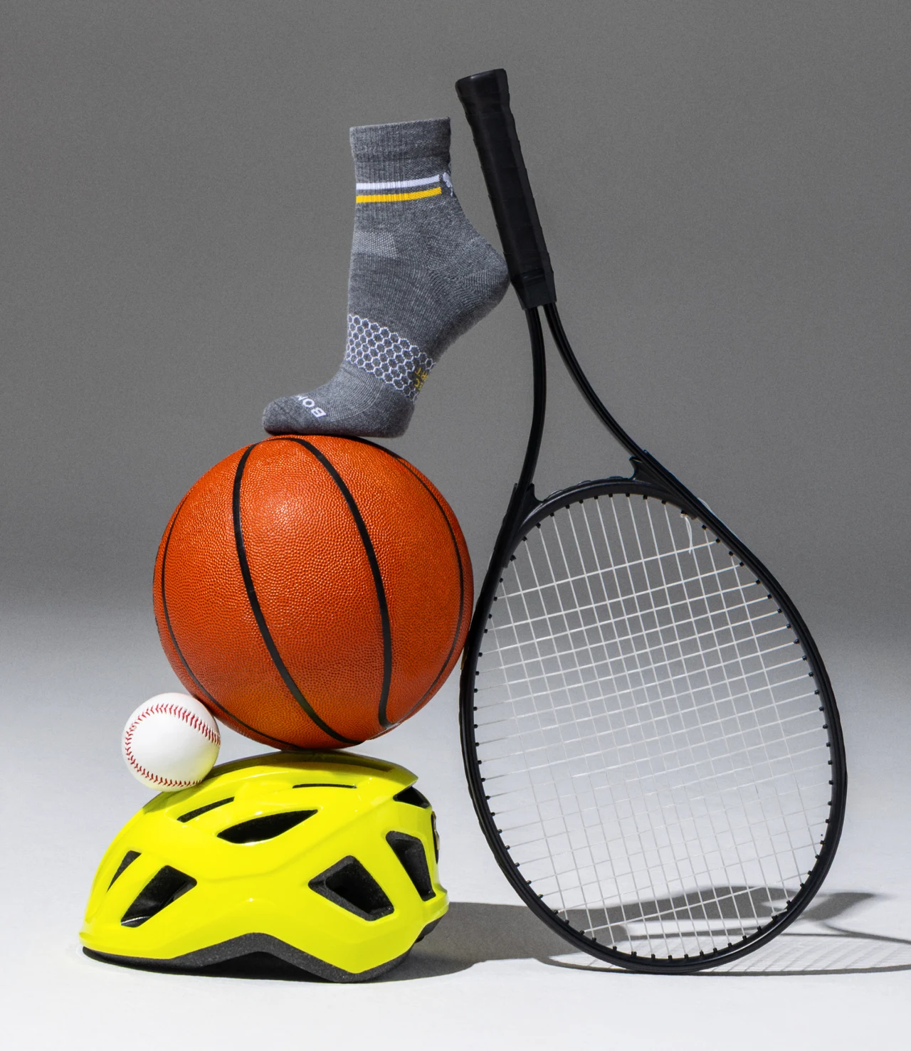 Still life of a grey athletic sock balanced on a basketball, a baseball and a neon yellow bike helmet, beside a black tennis racket on a grey backdrop.