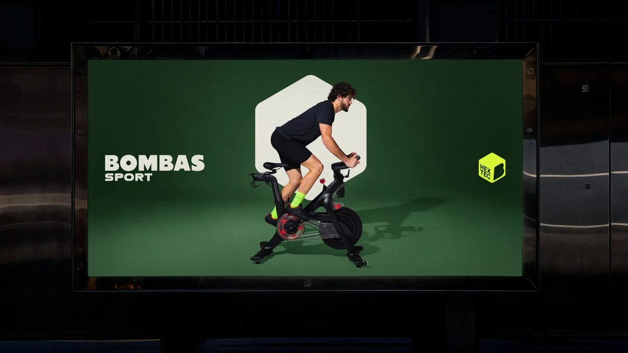 Backlit display in a dark space showing a green Bombas Sport ad: a man on a stationary bike inside a white hexagon and a neon Hex Tec badge.