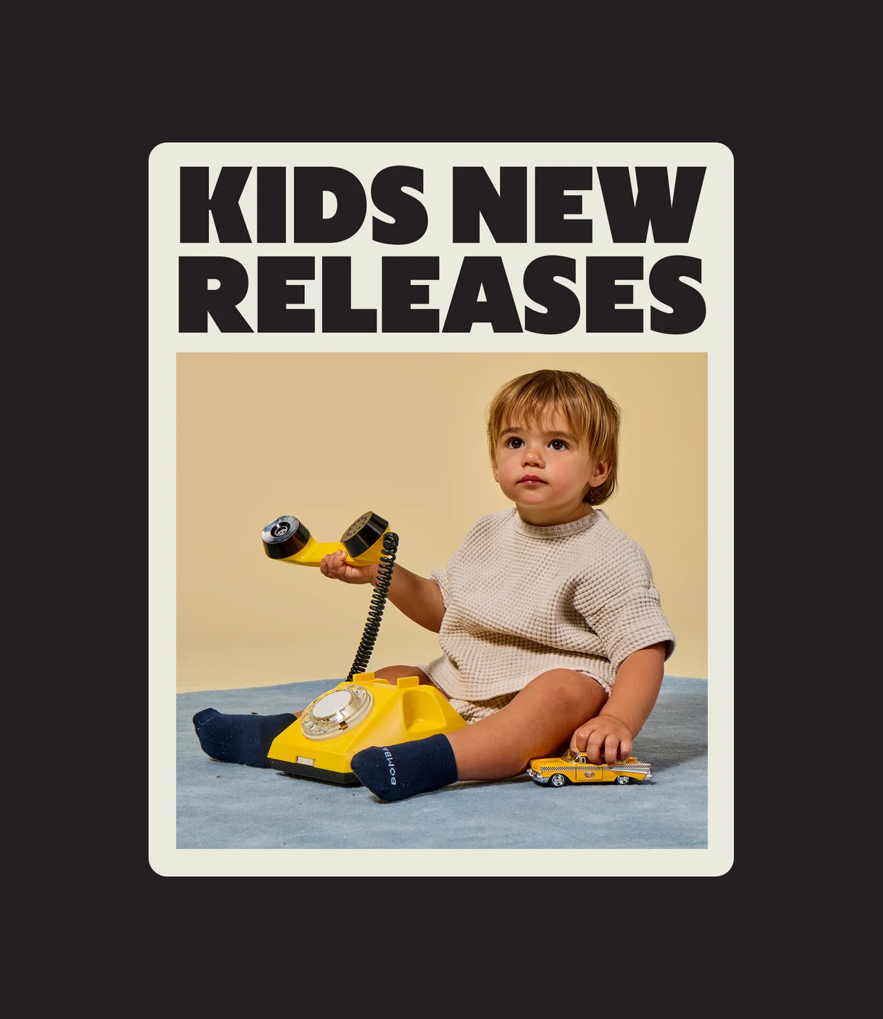 Kids New Releases card on a dark background, with a toddler in navy Bombas socks sitting on a blue rug with a yellow rotary phone and a toy taxi.