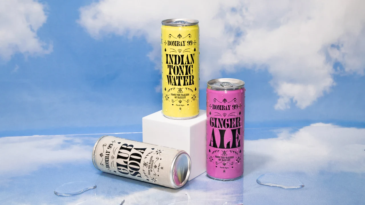 Yellow, pink and white Indian Tonic Water, Ginger Ale and Club Soda cans form a composition on a white pedestal against a cloudy sky.