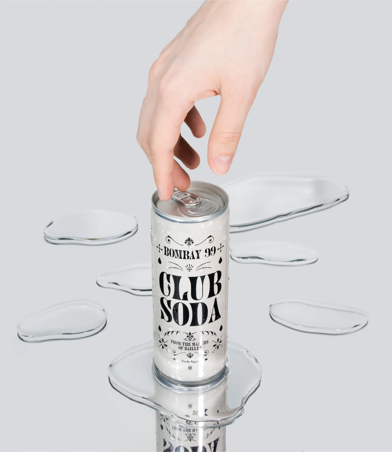 Vertical close-up of a hand opening a white Club Soda can, surrounded by small puddles and reflections on a pale gray surface.