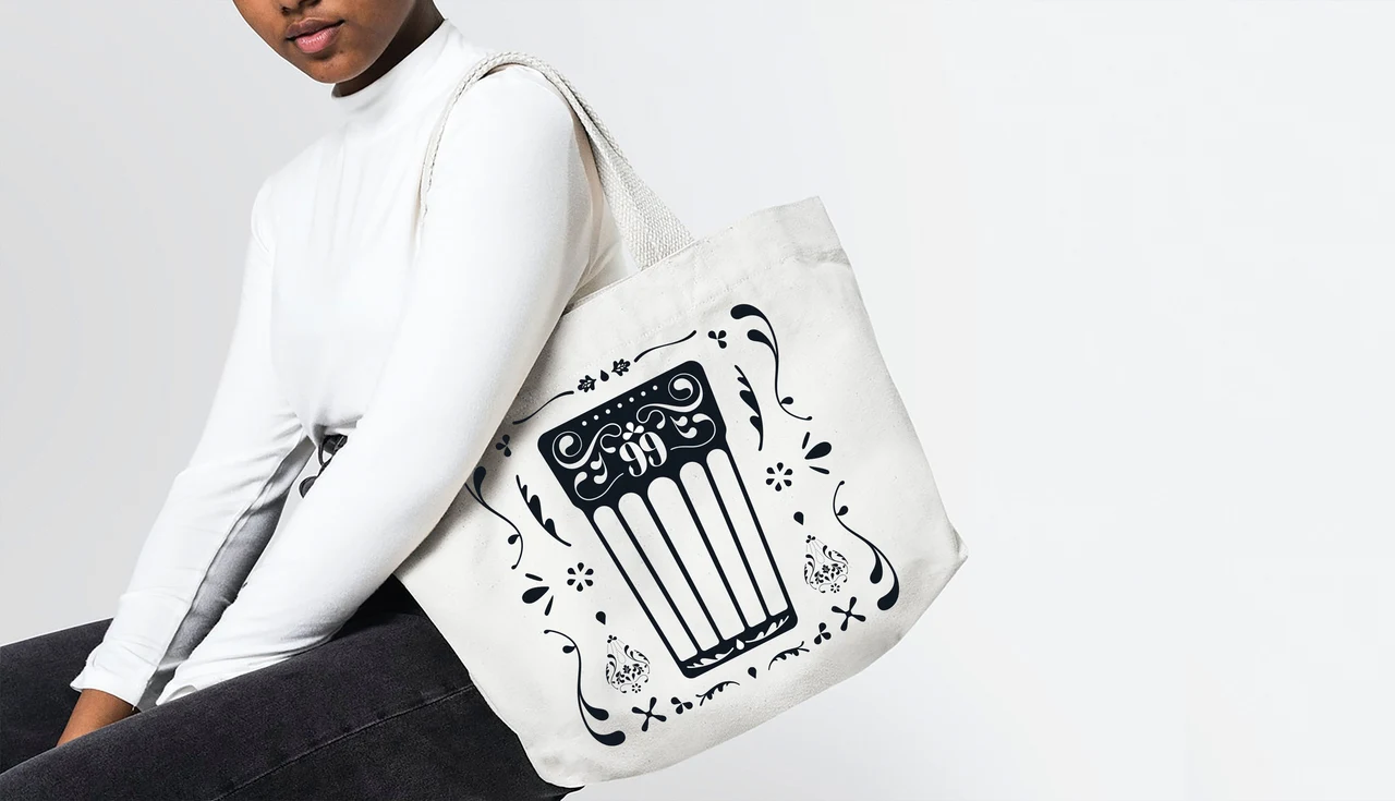 White tote bag on a person’s shoulder displays a large black tall-glass illustration surrounded by curves and small ornaments.