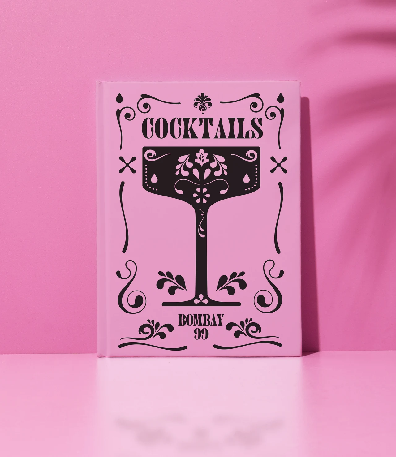 Upright pink cocktail menu shows a decorated black goblet, a Cocktails heading and a Bombay 99 signature on a pink table.