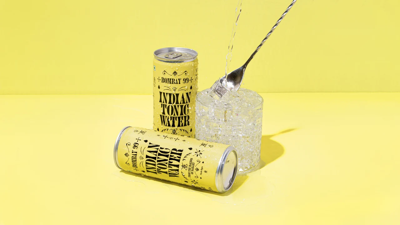 Two yellow Indian Tonic Water cans accompany an ice-filled glass and long spoon in a monochromatic yellow still life.