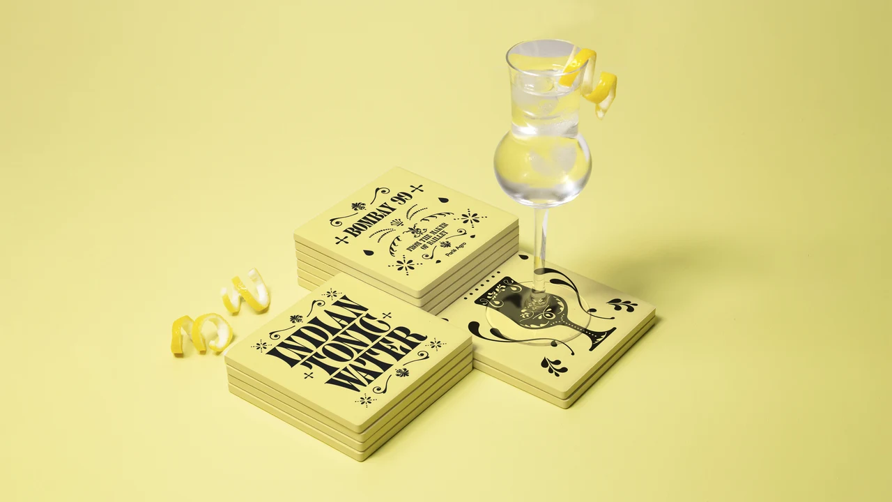Stacks of yellow printed materials with lettering and ornaments support a transparent stemmed glass, accompanied by citrus-peel spirals.