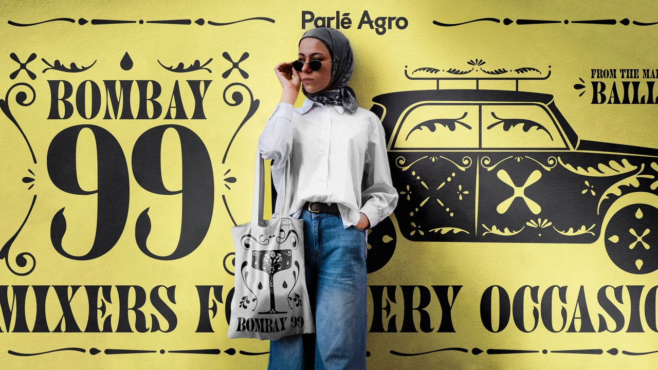 Person wearing a white shirt and jeans and carrying a tote poses before a large yellow Bombay 99 mural with black lettering and an ornamental car illustration.