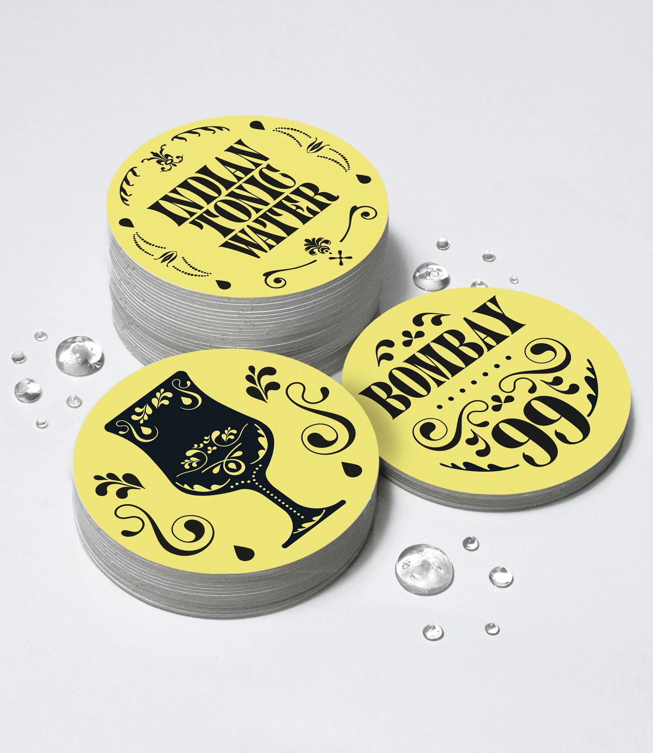 Stacks of circular yellow coasters feature Indian Tonic Water, Bombay 99 and a black ornamental goblet among transparent droplets.