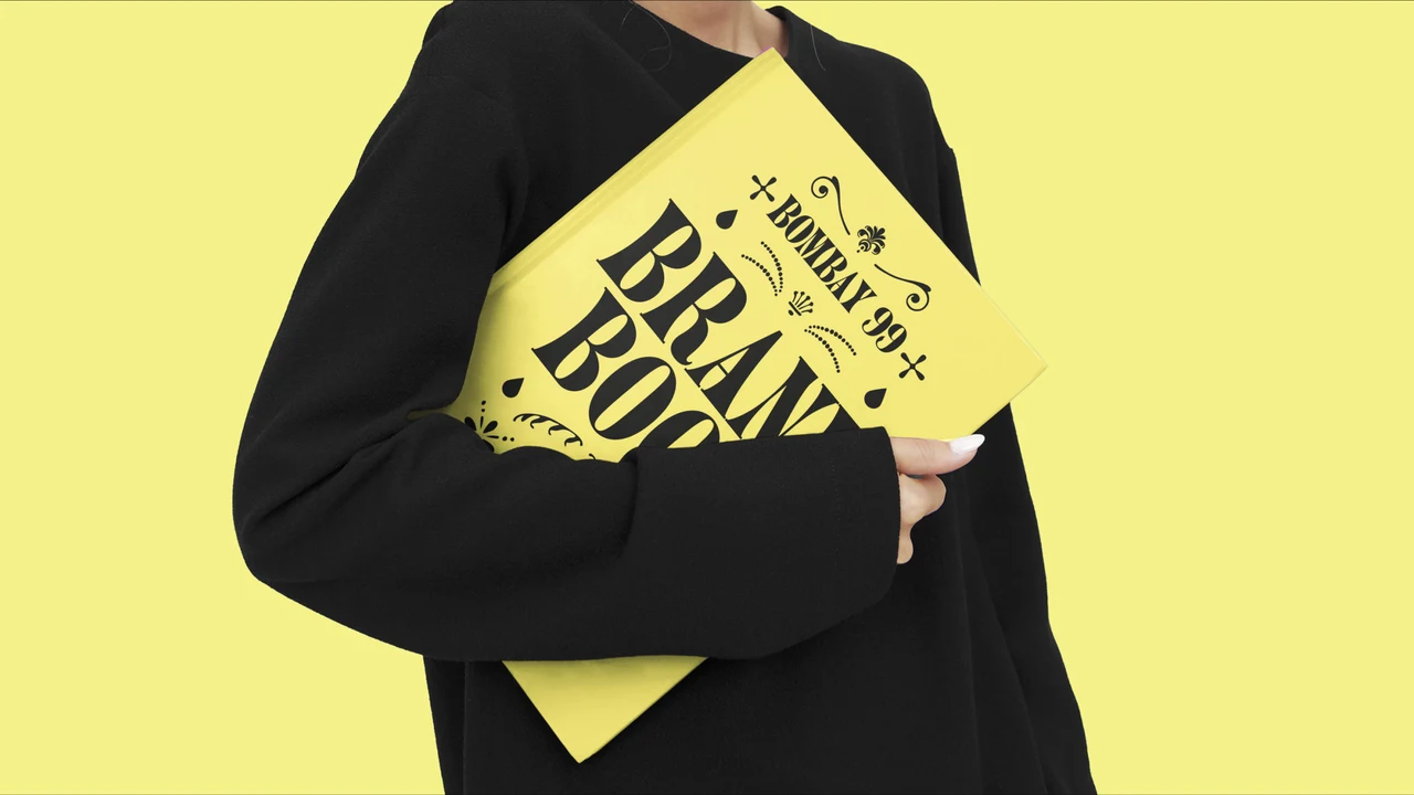 Person dressed in black holds a large yellow Bombay 99 Brand Book against their body, with expressive black typography covering its front.