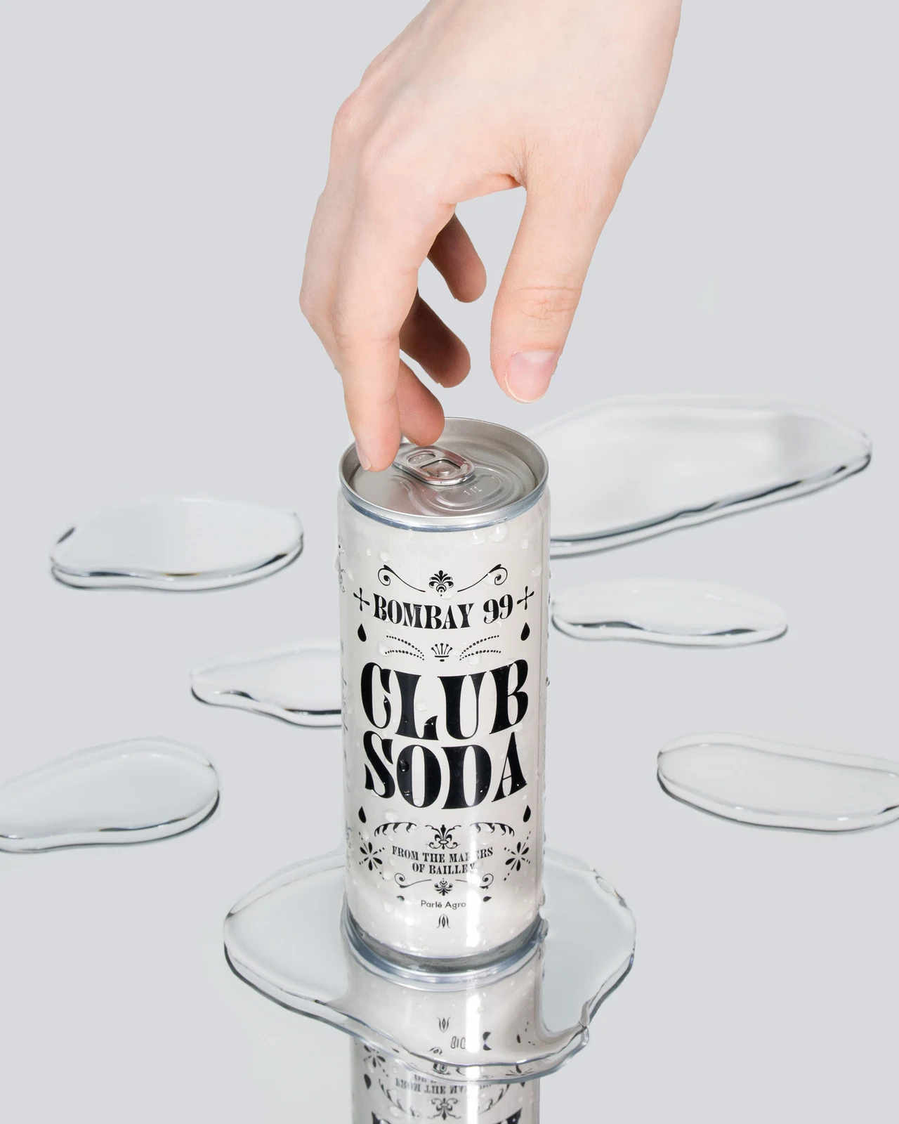 A hand opens a white Bombay 99 Club Soda can on a mirrored surface with transparent puddles, emphasizing black label lettering and ornaments.