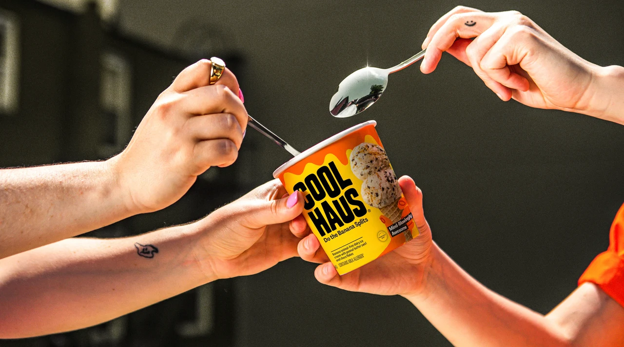 Hands share a yellow Coolhaus tub as two spoons approach, in sunlit photography against a dark blurred background.
