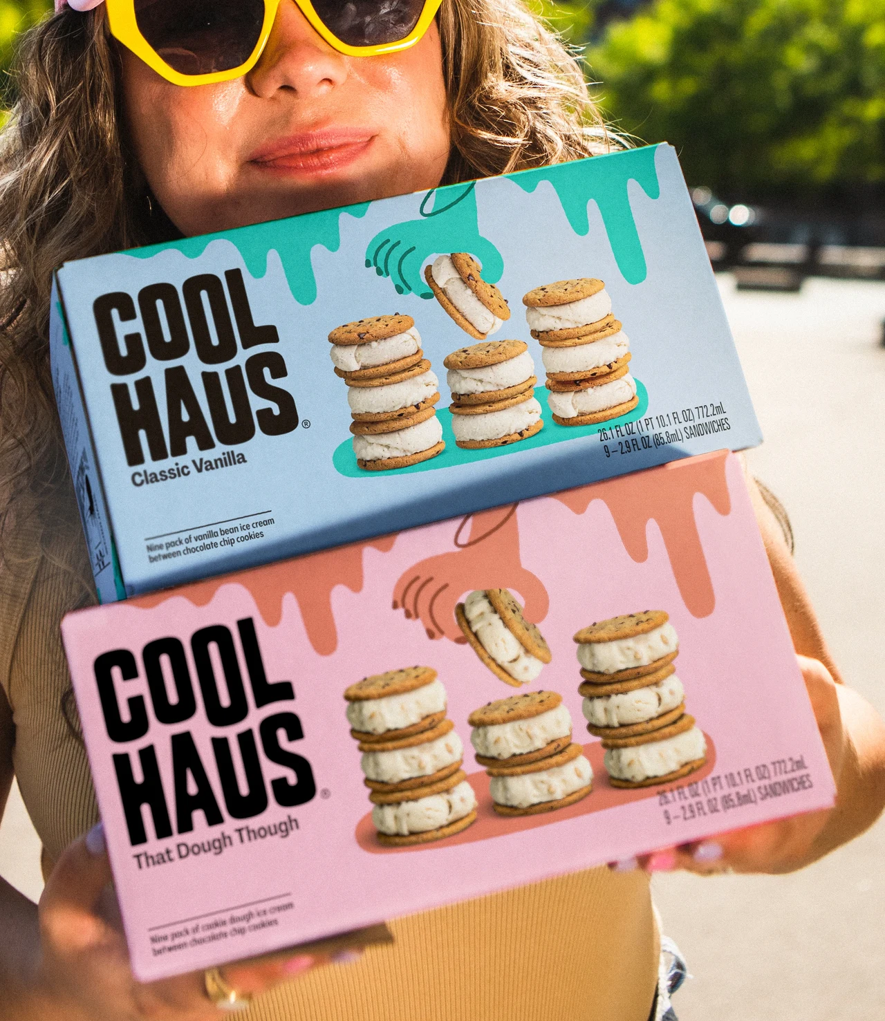 Person wearing yellow sunglasses holds two Coolhaus ice cream sandwich boxes, one blue and one pink, on a sunny street.