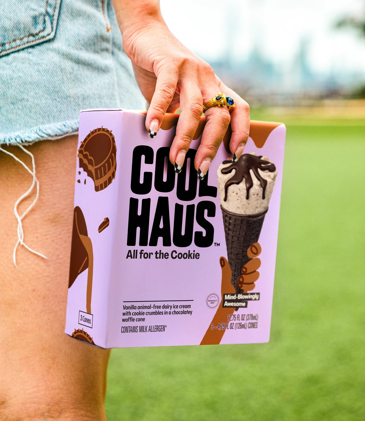 Close-up of a person in denim shorts holding a lilac Coolhaus All for the Cookie box with a photographed cone on its front, against blurred grass.