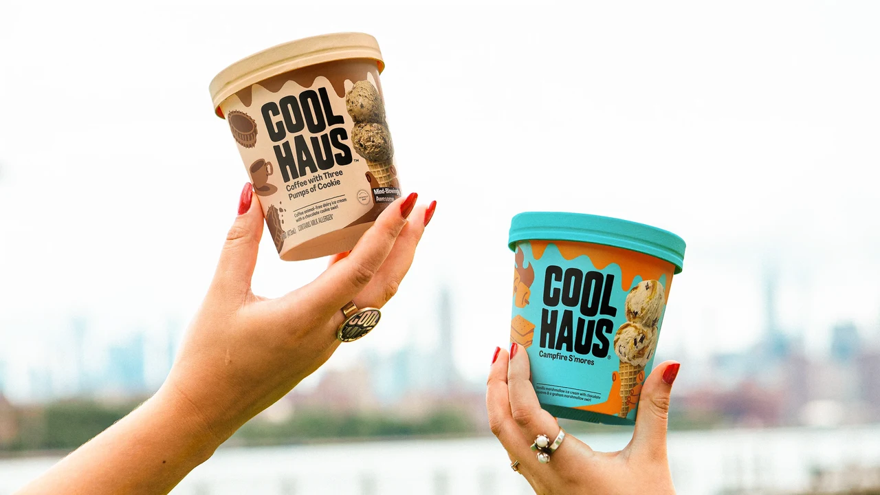 Two hands with red nails raise cream and blue Coolhaus tubs against a pale sky and a blurred cityscape.