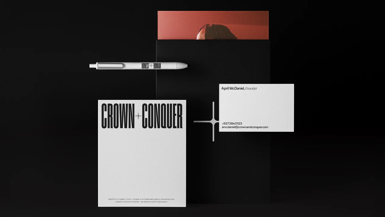 Black Crown + Conquer stationery: a notepad with the condensed CROWN + CONQUER wordmark, a founder's business card, a white Honest + Human pen and a large plus symbol.