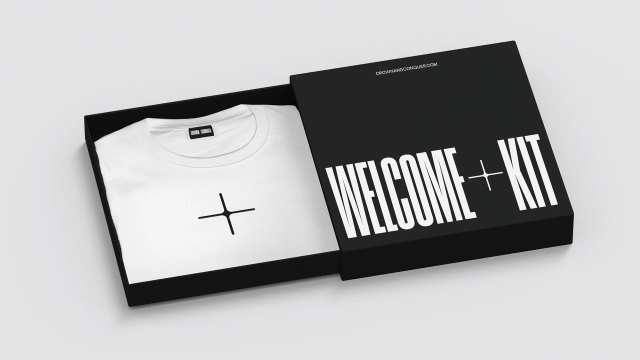 Black Welcome + Kit box with crownandconquer.com on the lid, opened to show a white T-shirt with a black plus symbol.