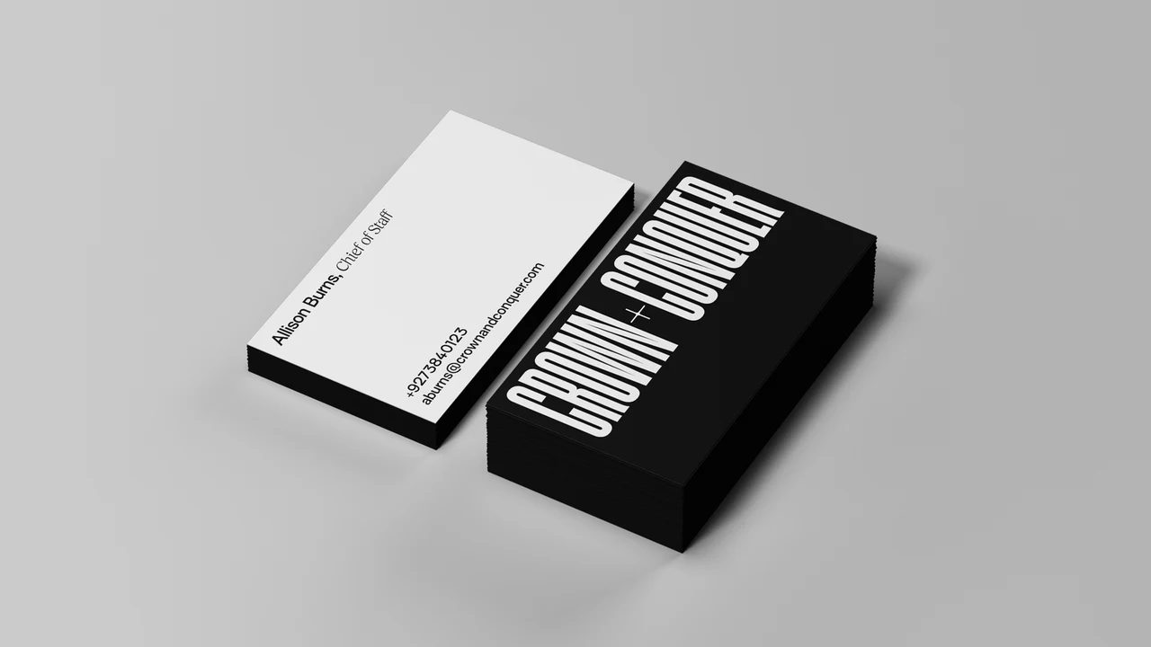 Two stacks of Crown + Conquer business cards on grey: black cards with the vertical CROWN + CONQUER wordmark and white cards with staff contact details.