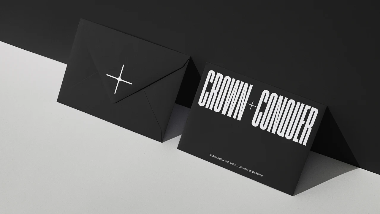 Black envelope with a white plus symbol and a folded black card with the CROWN + CONQUER wordmark and a Los Angeles address, against a black and white backdrop.