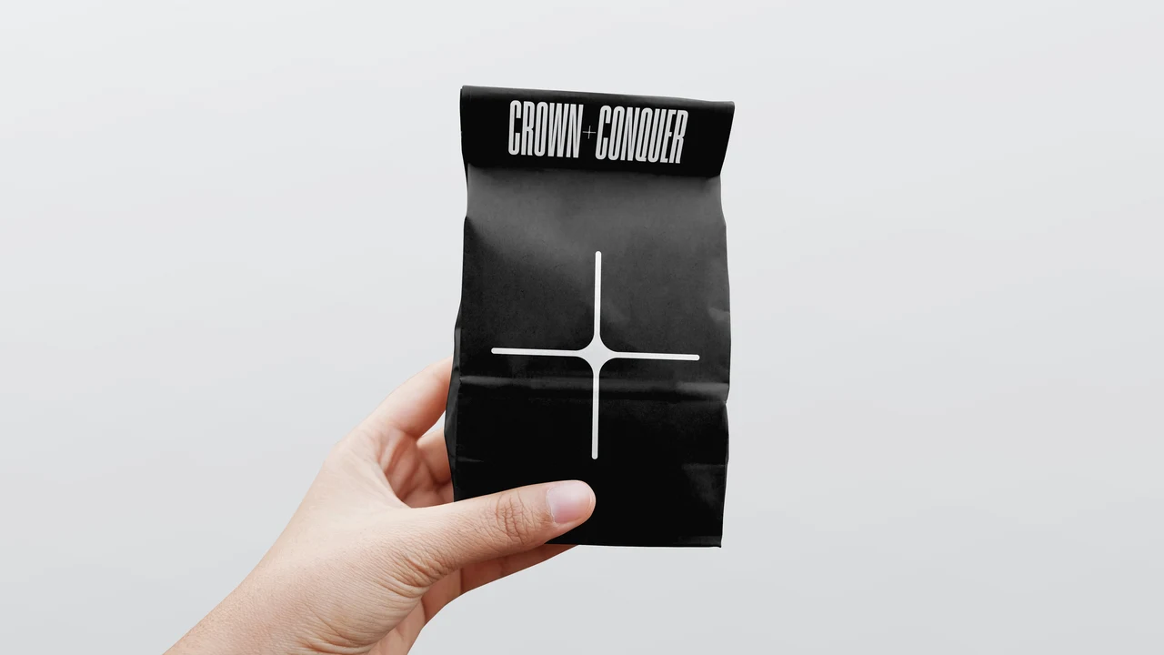 Hand holding a black paper bag with the CROWN + CONQUER wordmark at the top and a large white plus symbol, against a light grey background.