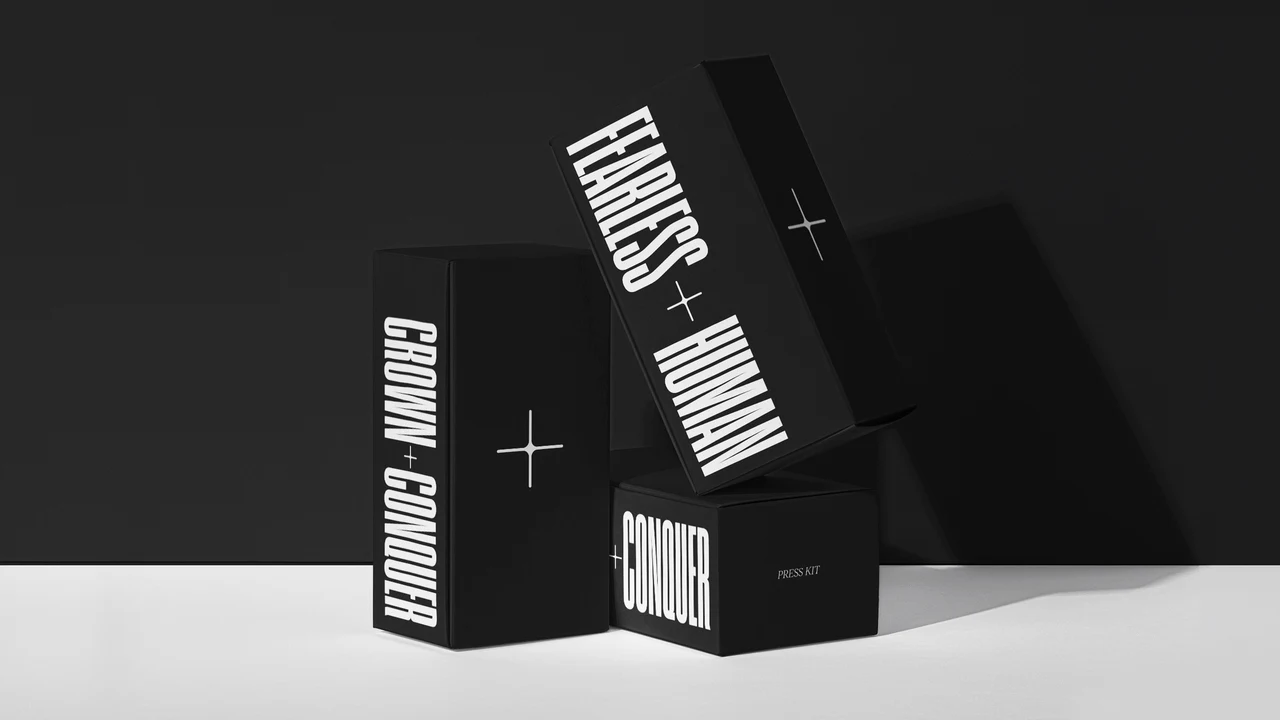 Three black boxes on a black and white set: a tall box with the vertical CROWN + CONQUER wordmark, a tilted Fearless + Human box and a small Press Kit box, each with a plus symbol.