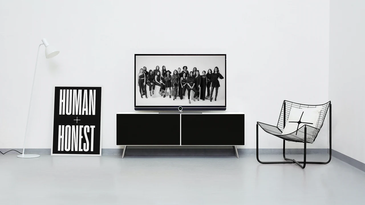 White room with a Human + Honest poster leaning on the wall, a floor lamp, a TV on a black console showing a black-and-white team photo and a wire chair with a plus-symbol cushion.