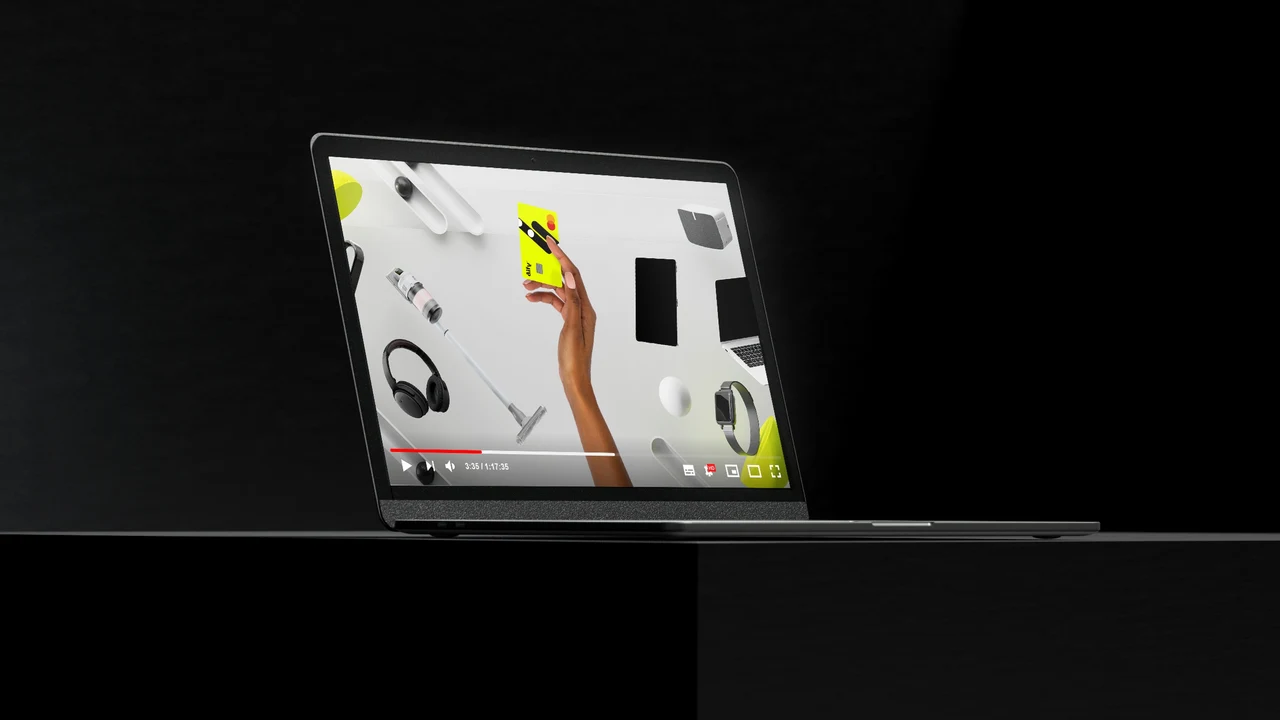 A laptop in a black setting plays a product film showing a hand lifting a yellow card among electronic devices.