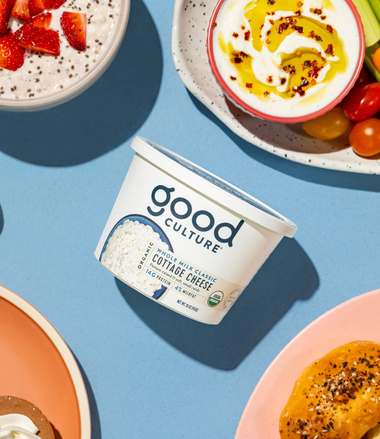 An overhead view shows a Good Culture tub on a blue background surrounded by dishes of cottage cheese with strawberries, olive oil and other toppings.