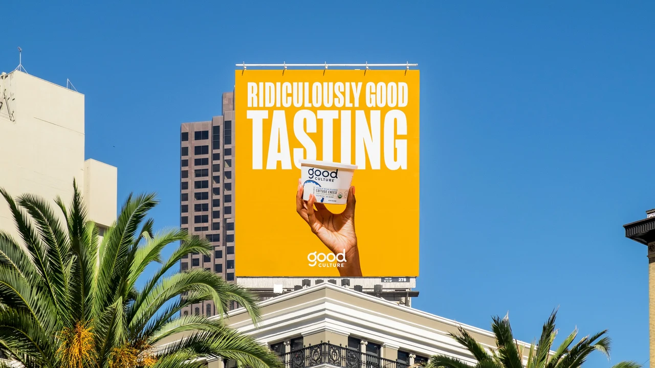 A vertical yellow billboard shows a hand raising a Good Culture tub beneath tall white lettering above a building and palm trees.