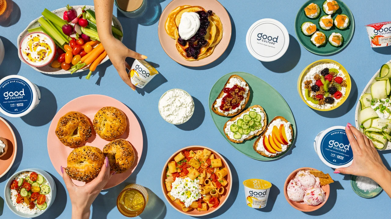 An overhead blue table holds varied dishes of fruit, salads, pancakes and cottage cheese, interspersed with Good Culture tubs and hands serving food.