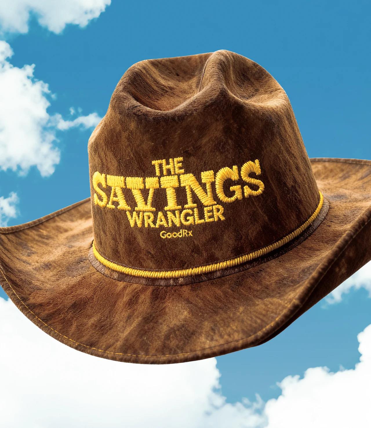 Brown suede cowboy hat floating in a blue sky with clouds, embroidered in yellow with The Savings Wrangler and GoodRx.