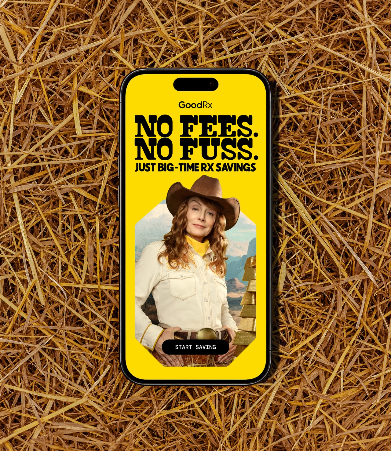 Smartphone lying on straw with a yellow GoodRx screen: No Fees. No Fuss. Just Big-Time Rx Savings, the cowgirl in a hat and a Start Saving button.