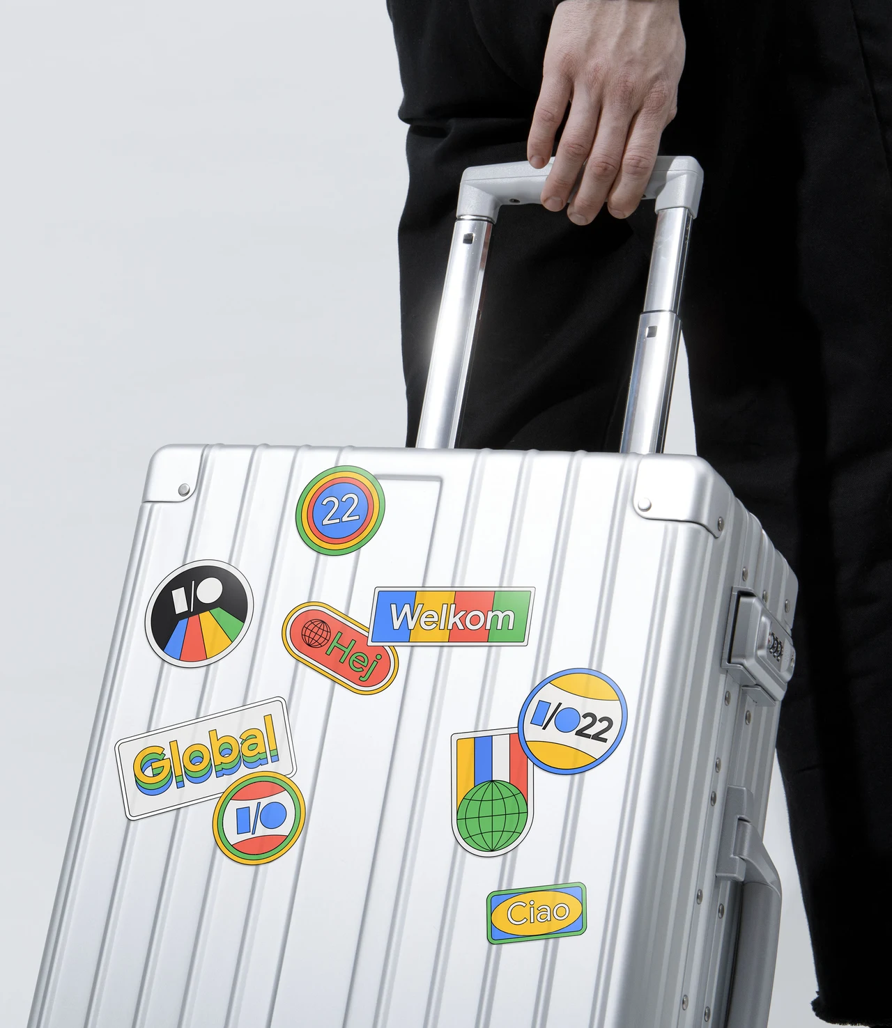 Mala rígida prateada é decorada com adesivos Google I/O 2022 de globos, logotipos e saudações em diferentes idiomas.