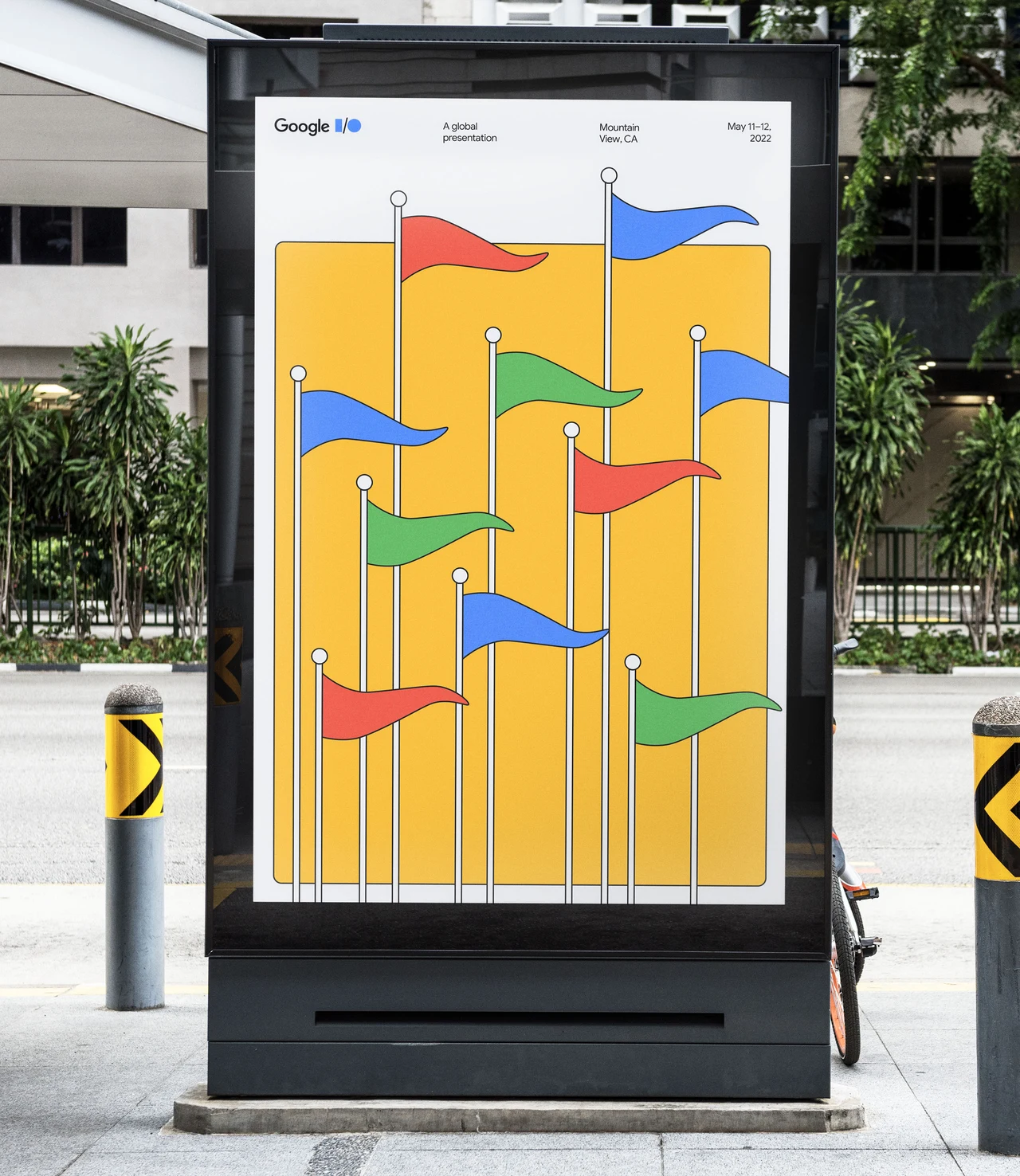 Totem publicitário exibe cartaz Google I/O com várias bandeiras azuis, vermelhas, amarelas e verdes em hastes finas sobre um grande campo amarelo.