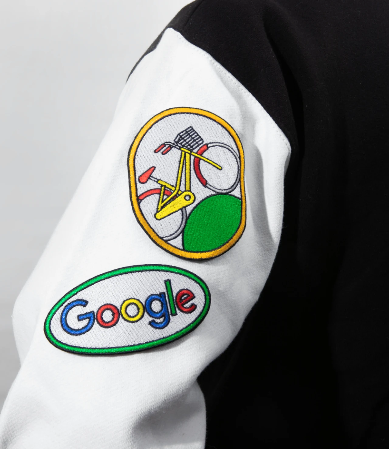 Close da manga branca de uma jaqueta mostra dois patches bordados: uma ilustração colorida com bicicleta e o logotipo Google.