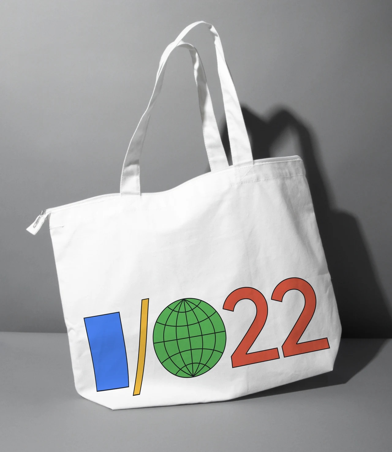 Ecobag branca com alças longas recebe a assinatura I/O 22 feita de retângulo azul, linha amarela, globo verde e números vermelhos.