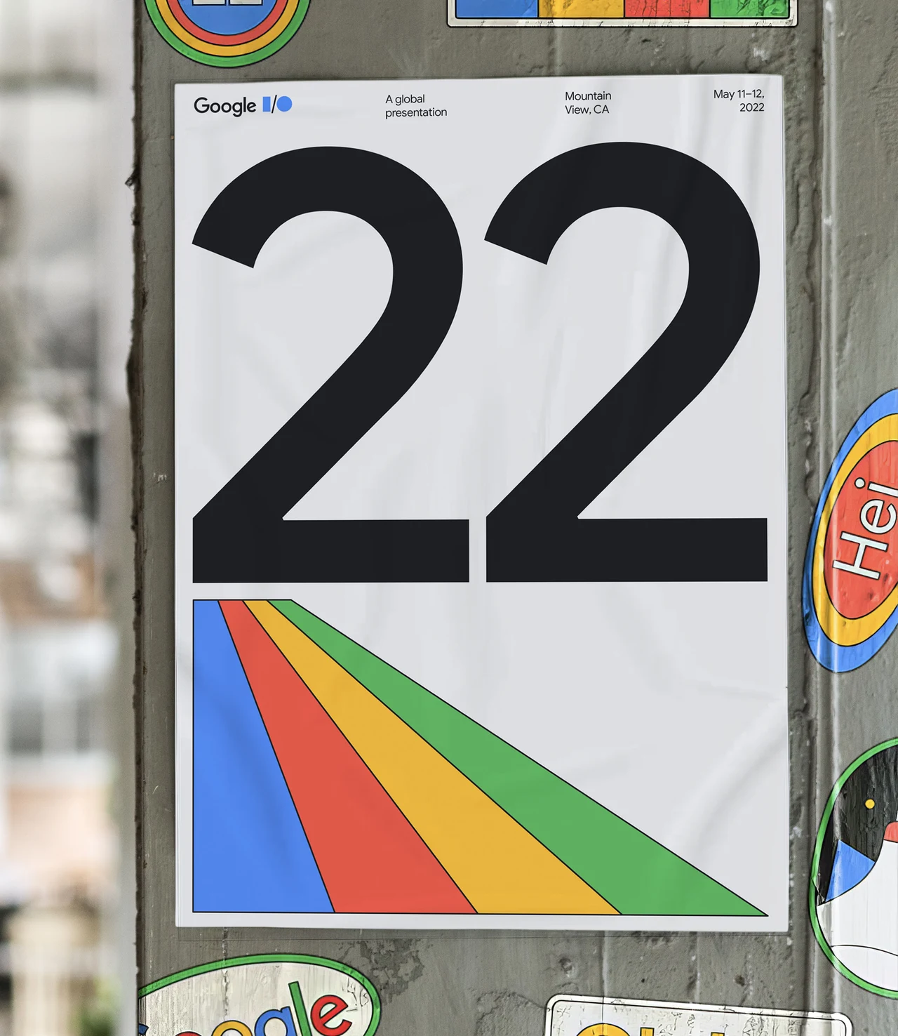 Cartaz colado em um poste destaca o número 22 em preto sobre branco e faixas diagonais nas cores Google, cercado por outros adesivos do evento.